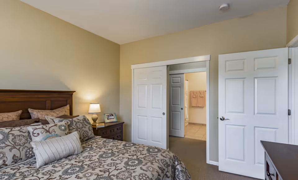 Bedroom with a patterned bedspread and wooden headboard, a nightstand with lamp, and open doors leading to a bathroom.