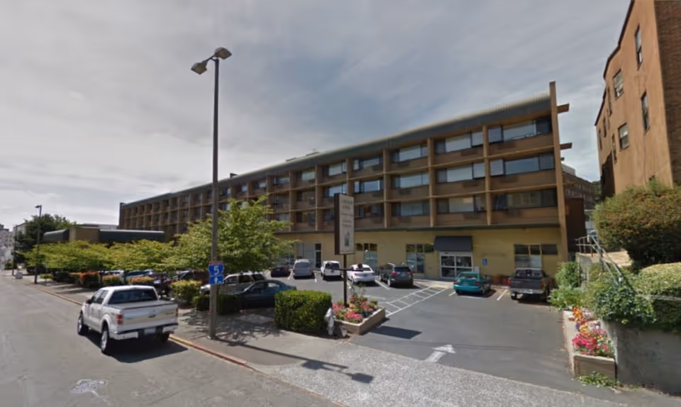 Exterior view of a multi-story senior living facility named Cascade Park Vista with a parking lot in front, several parked cars, trees, and a street with a white pickup truck driving by.
