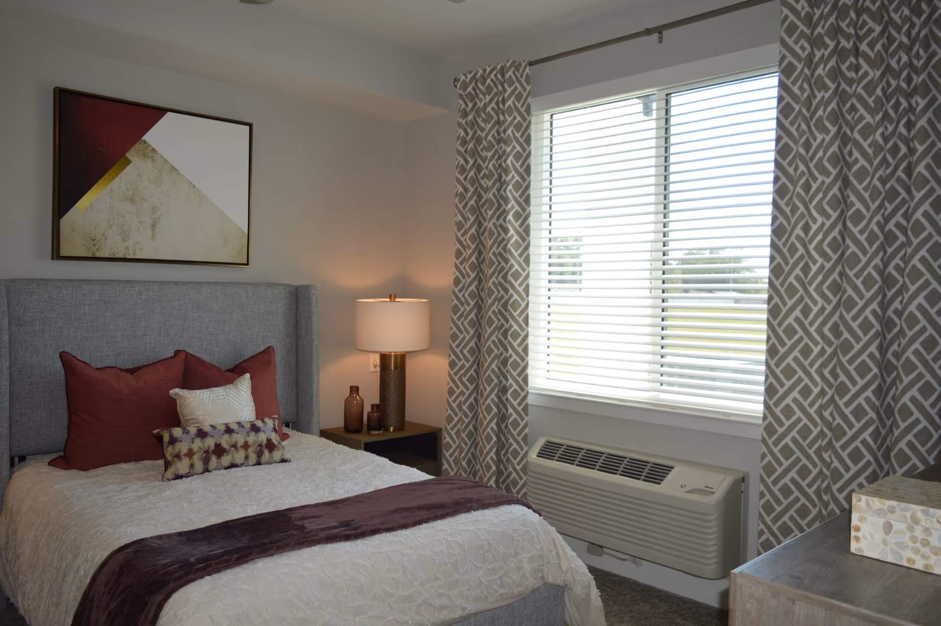 A bedroom with a made bed topped with decorative pillows, a bedside lamp and table, abstract wall art, and a window with blinds and patterned curtains.