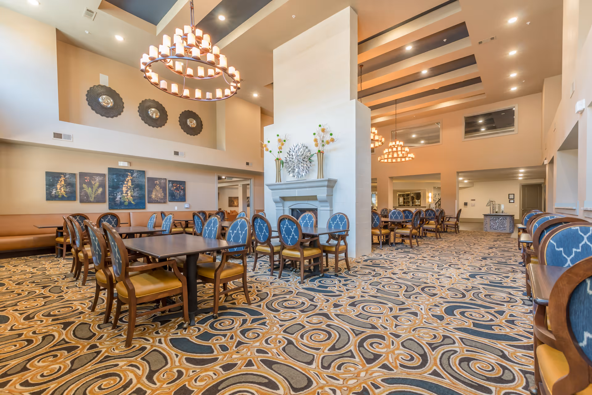 Spacious dining room with multiple wooden tables and chairs featuring blue patterned upholstery. The room has a high ceiling with modern chandeliers, decorative wall art, and a central fireplace adorned with vases and a metallic wall sculpture. The carpet has an intricate swirling pattern in shades of blue, beige, and brown.