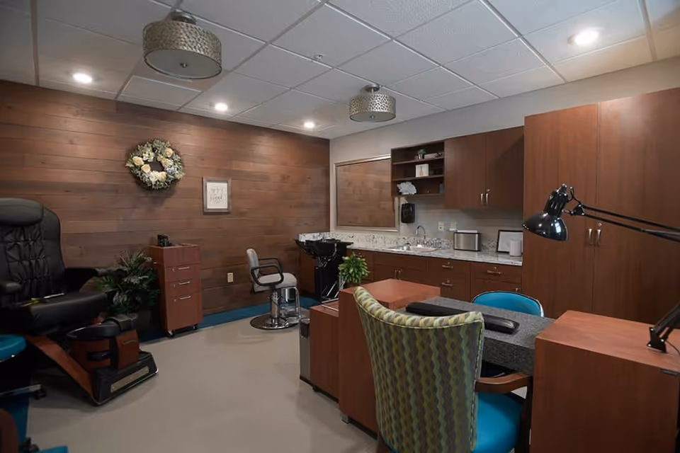 Interior view of a salon area in a senior living facility with a pedicure chair, a hair washing station, a styling chair, wooden cabinets, a countertop with a sink, and a desk with a patterned chair and a blue chair. The walls are wood-paneled and decorated with a floral wreath and a framed picture.