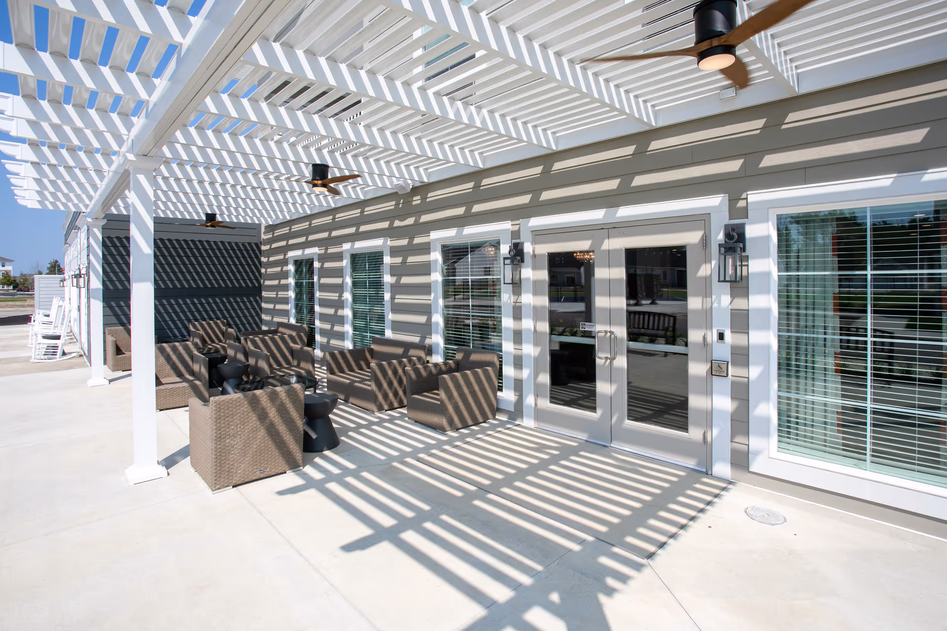 Outdoor patio area at Provision Living at Findlay with a white pergola casting striped shadows on the concrete floor. The patio features several brown wicker chairs with small black side tables arranged in seating groups. Ceiling fans are mounted under the pergola, and there are large windows and glass double doors leading inside the building.