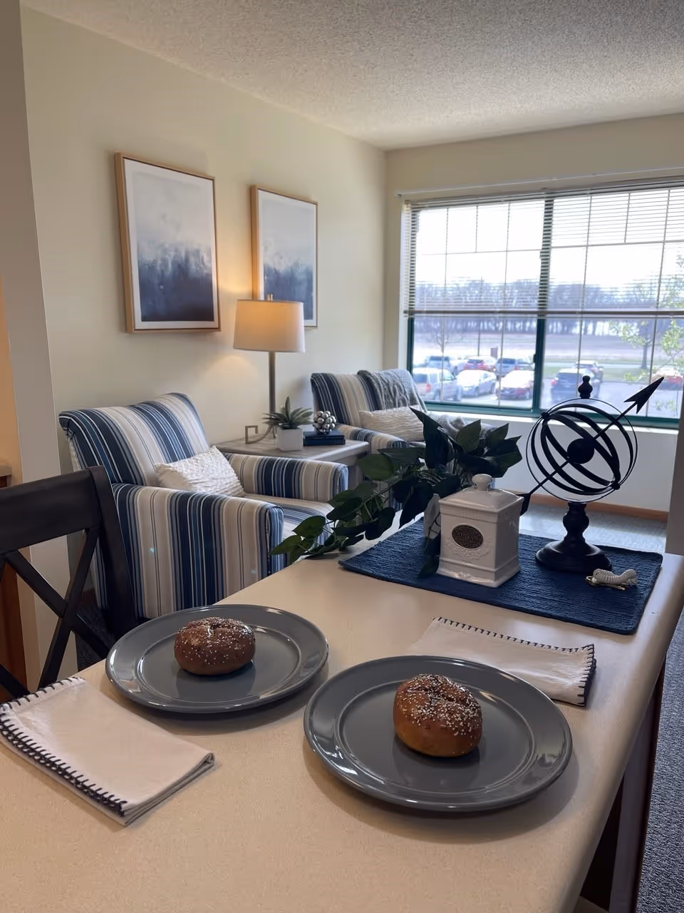 Cozy living area with two striped armchairs by a large window and a countertop set with two plates each holding a bagel.