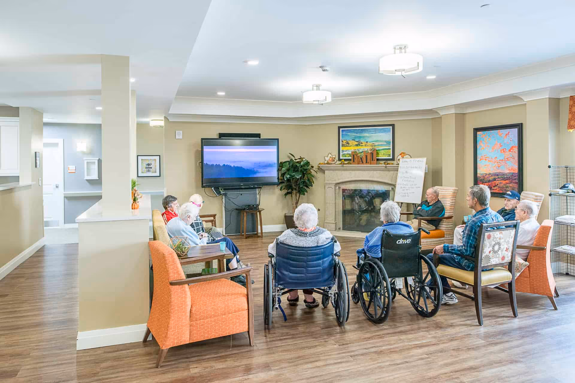 A group of elderly people sitting in a circle in a well-lit common room with wooden flooring. Some are seated in wheelchairs while others are in armchairs. A flat-screen TV is mounted on the wall, and there is a fireplace with framed artwork above it. The room has beige walls and modern ceiling lights.