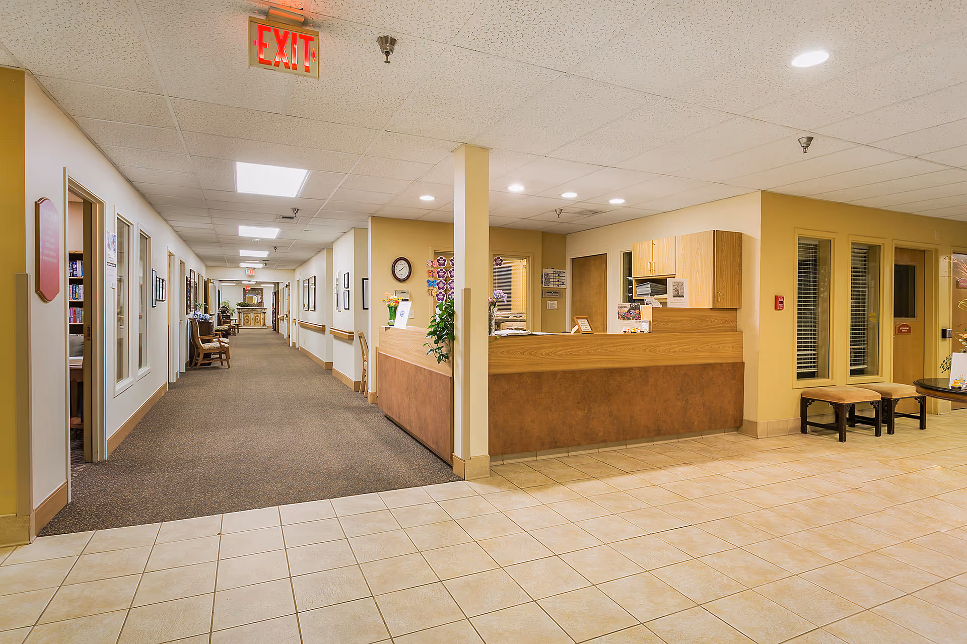 Interior view of a senior living facility hallway with a reception desk on the right side. The hallway has carpeted flooring and is lined with doors and windows on the left. The reception area has a wooden counter, cabinets, and some chairs nearby. The ceiling has recessed lighting and an illuminated exit sign is visible.