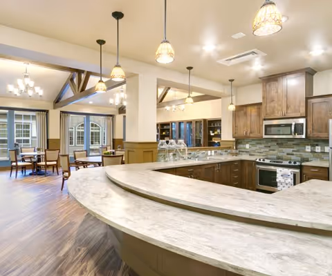 Open communal kitchen and dining area with a curved stone countertop, pendant lights, wooden cabinets, and dining tables in the background.