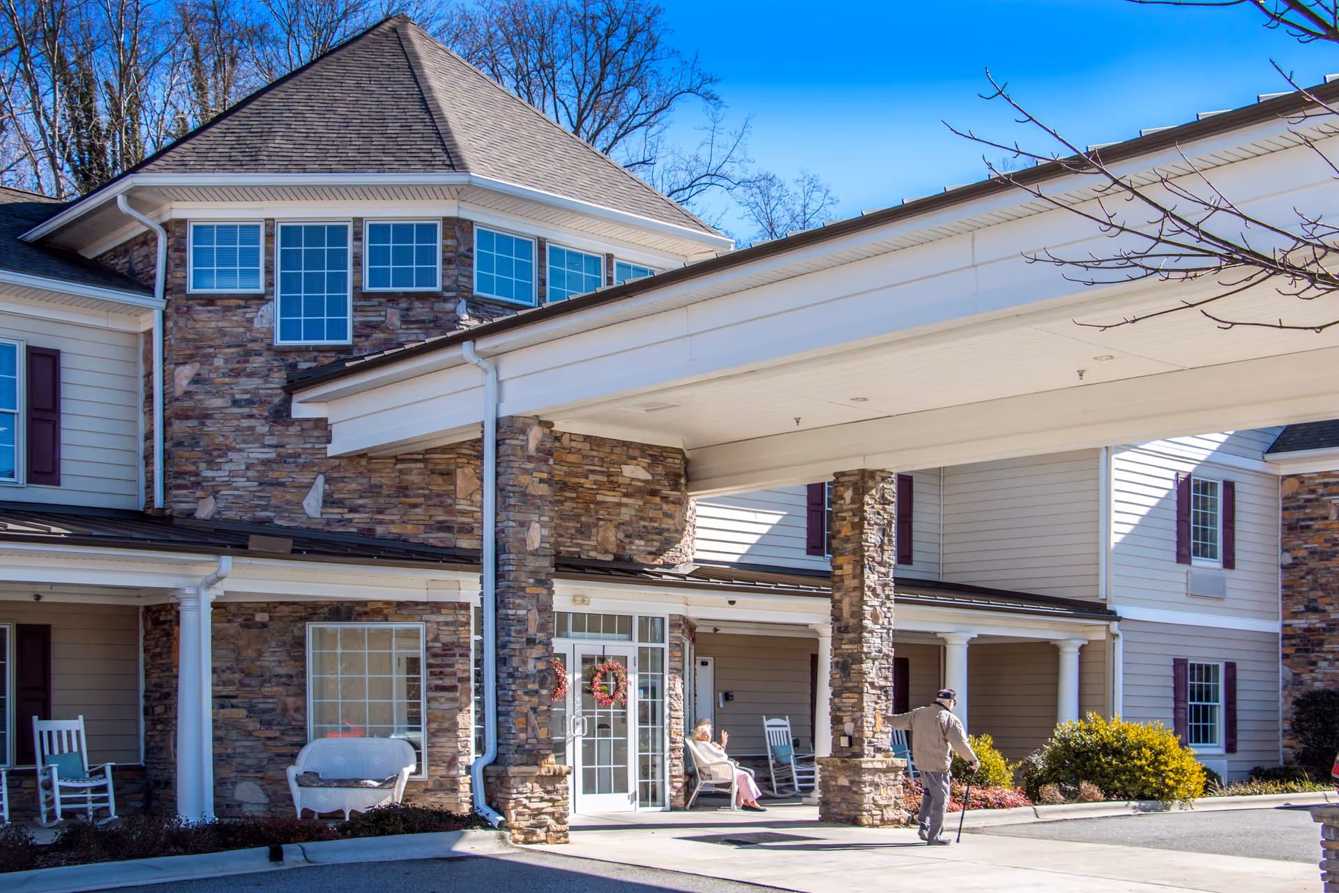 Exterior view of Arbor Ridge At Kernersville senior living facility showing a covered entrance supported by stone pillars. A person with a cane is walking near the entrance, and another person is sitting on a rocking chair on the porch. The building has stone and siding exterior with multiple windows and a clear blue sky above.