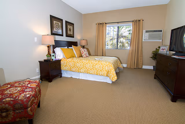 Spacious bedroom with a bed in yellow patterned bedding, bedside tables with lamps, a dresser with a TV, and a window with beige curtains.