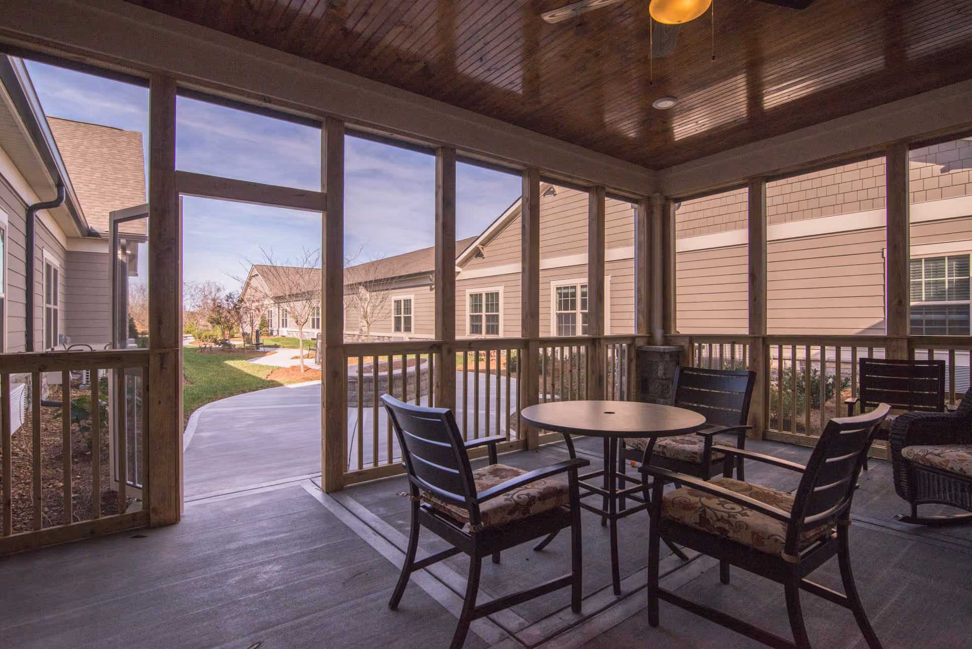 A screened-in porch area with a round table and four cushioned chairs. The porch has wooden railings and a wooden ceiling with a ceiling fan. Outside the porch, there is a paved walkway and beige buildings with windows under a clear sky.