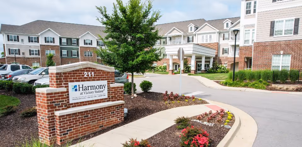 Exterior view of Harmony at Victory Station senior living facility showing a brick sign with the facility name, a landscaped garden with flowers and shrubs, a driveway, parked cars, and a multi-story building with white siding and brick accents under a partly cloudy sky.