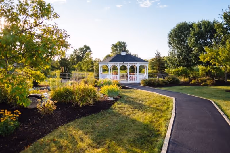A paved pathway leads through a well-maintained garden with various green shrubs and trees towards a white gazebo in the distance under a clear sky.