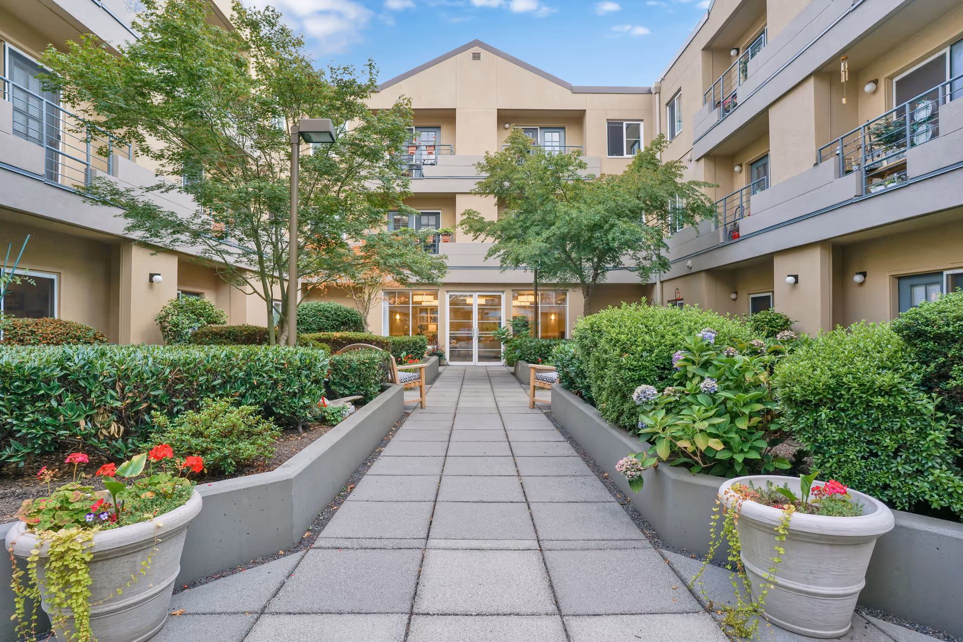 Outdoor courtyard area of Island House Assisted Living featuring a paved walkway lined with green shrubs, flowering plants in large pots, and trees. The courtyard is surrounded by a beige multi-story building with balconies and windows, under a partly cloudy blue sky.
