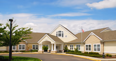 Exterior view of a single-story senior living facility building with beige siding, white trim, and a peaked roof under a partly cloudy blue sky. The entrance features a covered portico with columns, and there is a flagpole with an American flag near the entrance. Well-maintained landscaping with green grass, shrubs, and a tree is visible in front of the building.
