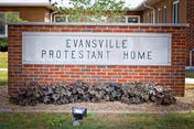 A brick sign with a white rectangular plaque displaying the text 'EVANSVILLE PROTESTANT HOME' in capital letters, surrounded by small plants and grass, with a residential building visible in the background.