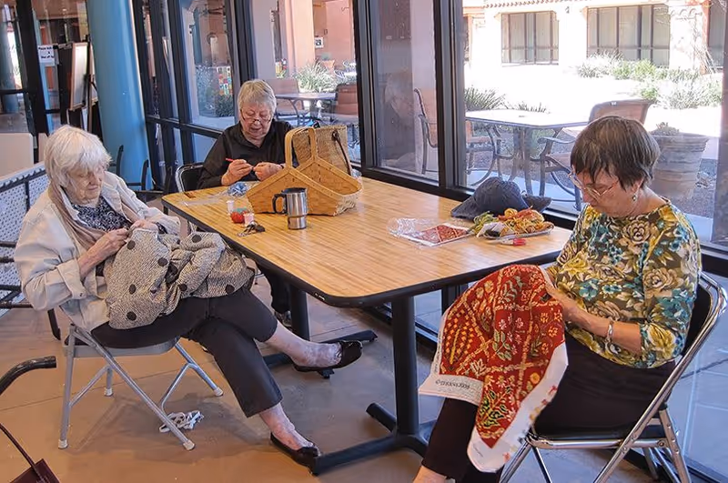 Three elderly women sitting around a rectangular wooden table near large windows, engaged in sewing or knitting activities. The room has a bright and airy atmosphere with outdoor patio furniture visible through the windows.