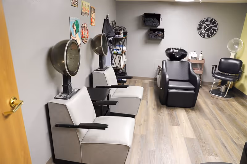 Interior view of a hair salon area with two vintage hair dryers attached to beige chairs on the left, a black shampoo chair with a sink in the center, a black salon chair on the right, a wall clock, and shelves with towels and hair products.