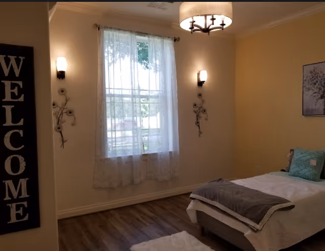 A cozy, softly lit bedroom with a single bed, window with sheer curtains, wall sconces, and a vertical 'WELCOME' sign.