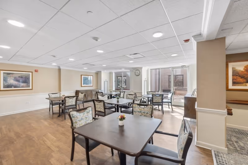 A bright dining room with several square tables and cushioned chairs arranged neatly. The room has wood flooring, beige walls with white trim, and framed landscape paintings. Large windows allow natural light to fill the space, and a clock is mounted on the wall near the windows.