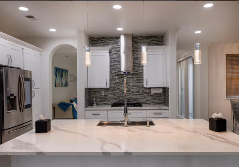 Modern kitchen with a large white marble island countertop featuring a built-in sink and faucet. Stainless steel refrigerator on the left, white cabinets, and a tiled backsplash behind a stainless steel range hood and stove. Pendant lights hang above the island, and a glimpse of a living area with a couch and artwork is visible through an archway.