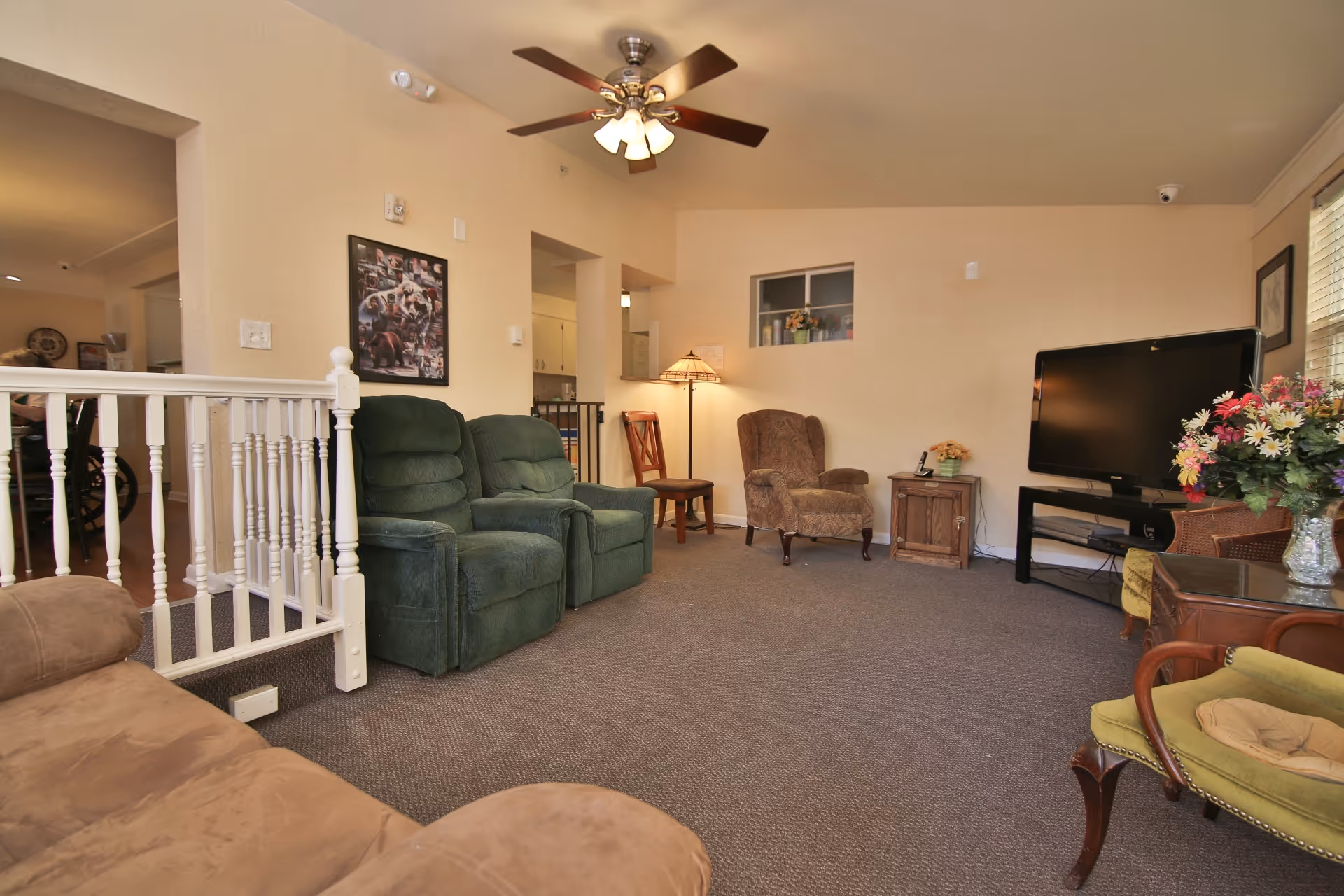 A cozy living room with beige walls and carpeted floor featuring two green recliner chairs, a brown armchair, a wooden side table with a lamp, a flat-screen TV on a black stand, and a vase of colorful flowers on a wooden table. There is a ceiling fan with lights and a white railing separating the living room from an adjacent area.