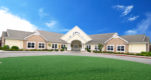 Front entrance of a single-story senior living building with a covered porte-cochère, circular driveway and a well-kept lawn under a blue sky.