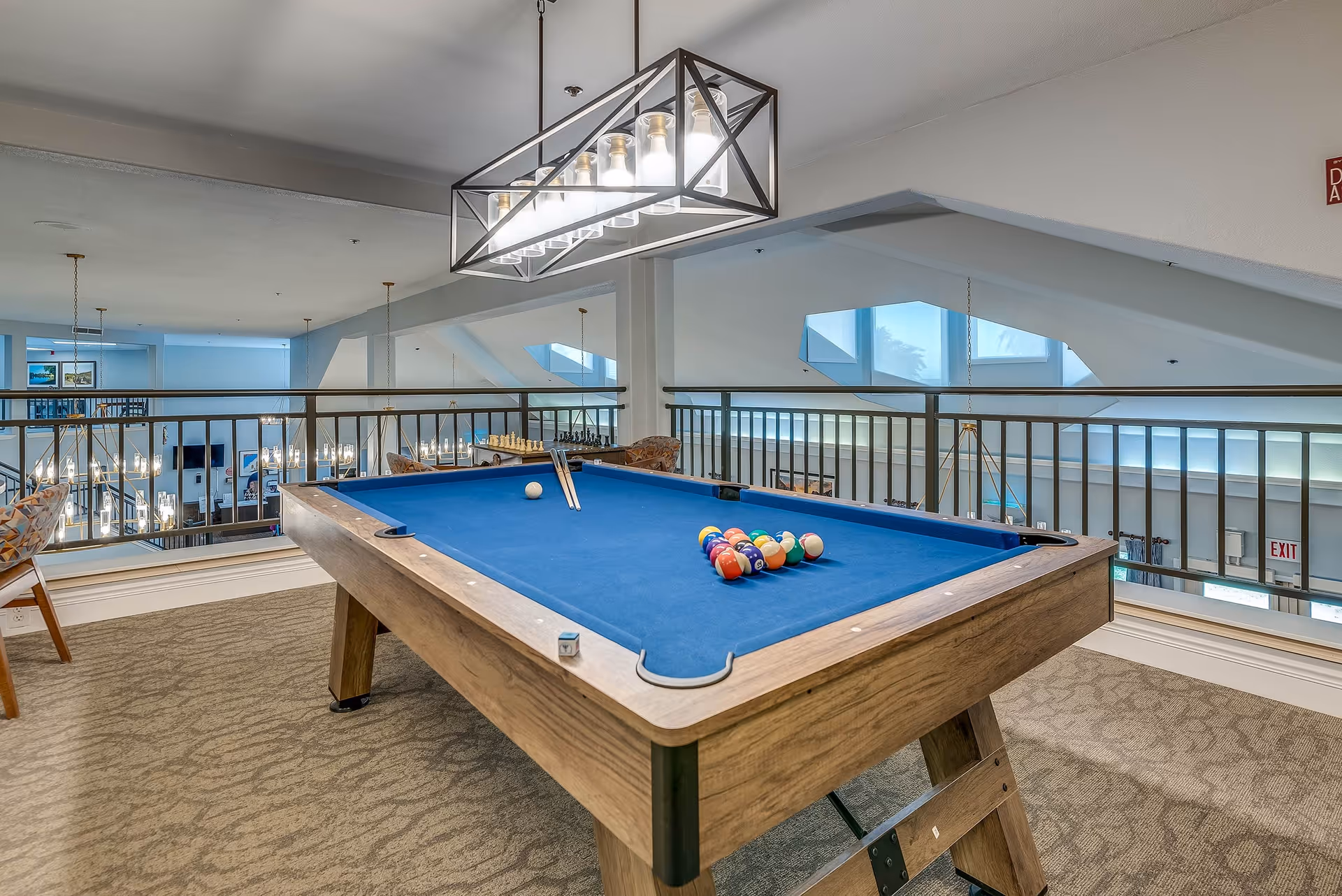 A blue-felt pool table in a bright indoor lounge overlooking an atrium with railings and pendant lights.