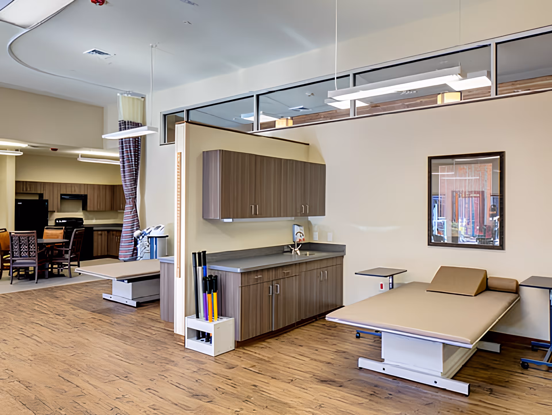 A spacious room with two adjustable medical examination tables, a countertop with cabinets above and below, a sink, and a set of colorful exercise sticks in a holder. The room has wood flooring and neutral-colored walls. In the background, there is a dining area with a table and chairs, a refrigerator, and kitchen cabinets.