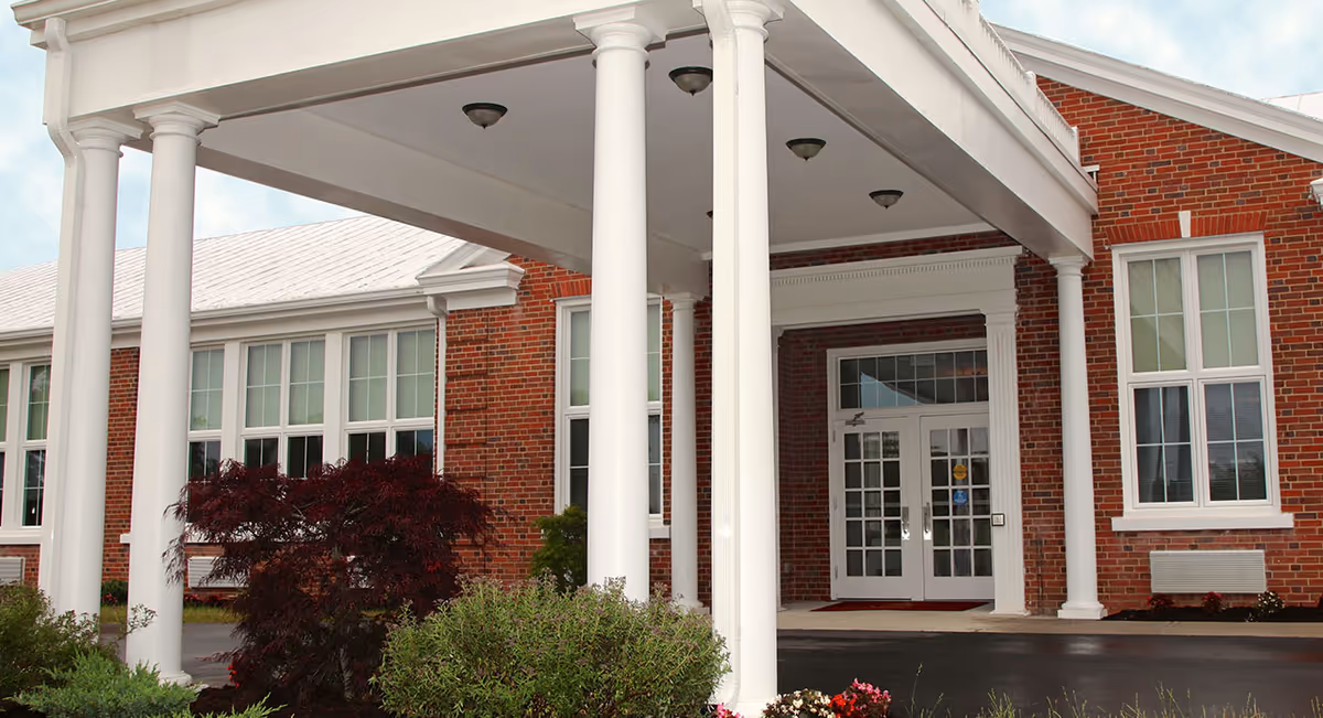 Entrance of a senior living facility with white columns supporting a covered driveway. The building is made of red brick with large windows and white trim. There are bushes and flowers planted near the entrance.