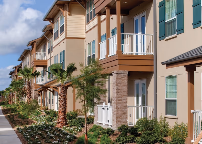 Exterior view of a multi-story residential building with balconies, palm trees, and landscaped greenery along a sidewalk under a partly cloudy sky.