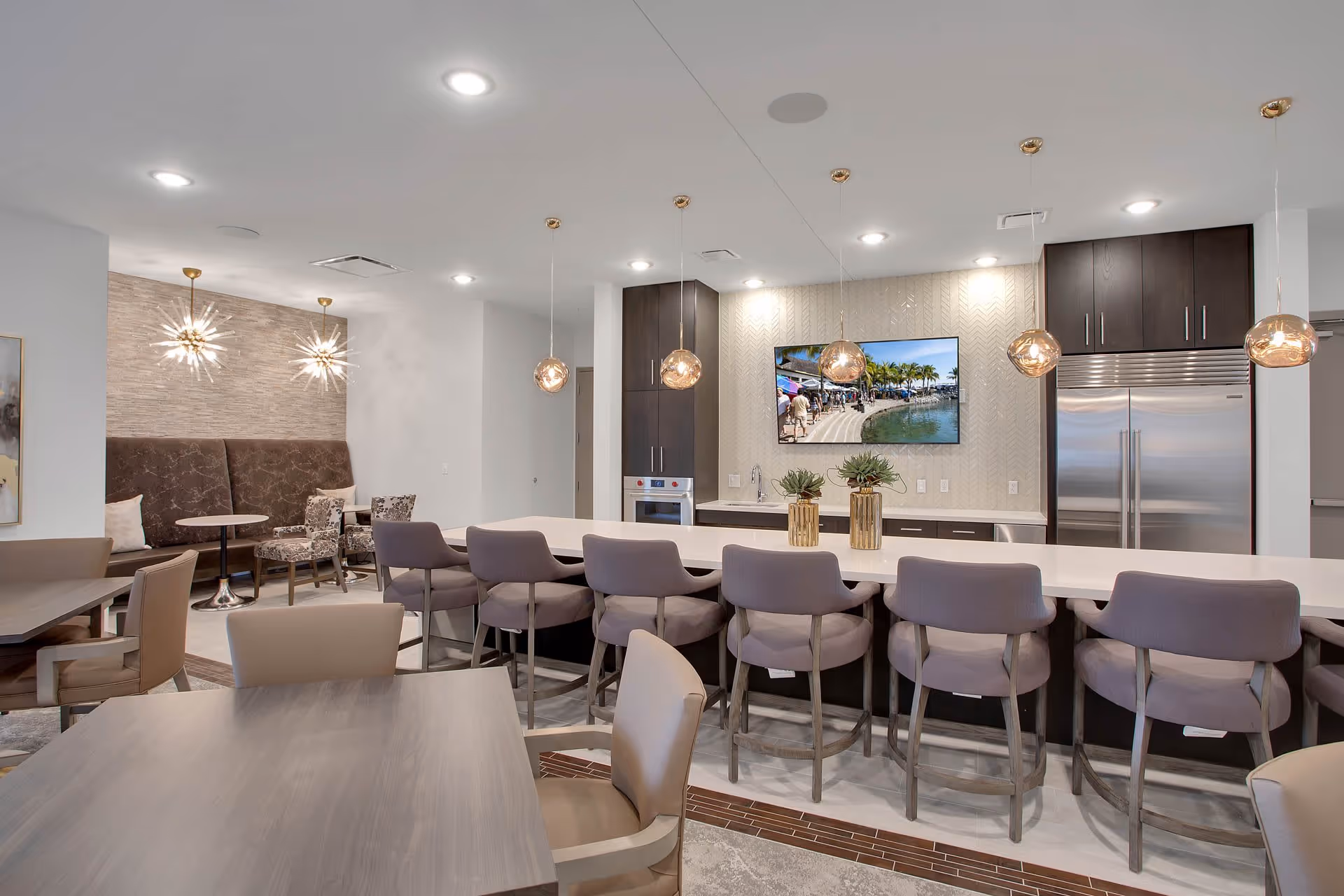 Modern communal kitchen and dining area with a long island, bar stools, pendant lights, tables and a wall-mounted TV.