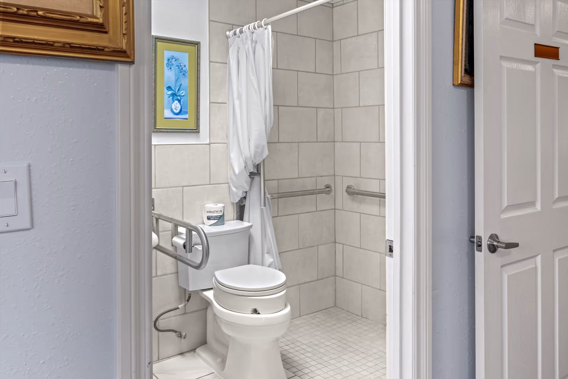 View into a bathroom featuring a white toilet with a grab bar on the left side, a tiled walk-in shower with grab bars on two walls, a white shower curtain, and a framed floral picture on the wall.