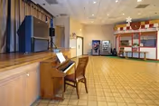 A large multipurpose room with a wooden piano and chair in the foreground on the left side. The room has a tiled floor, a stage area with curtains, and a concession stand or snack bar area on the right side. The ceiling has recessed lighting, and there are vending machines visible in the background near the concession stand.