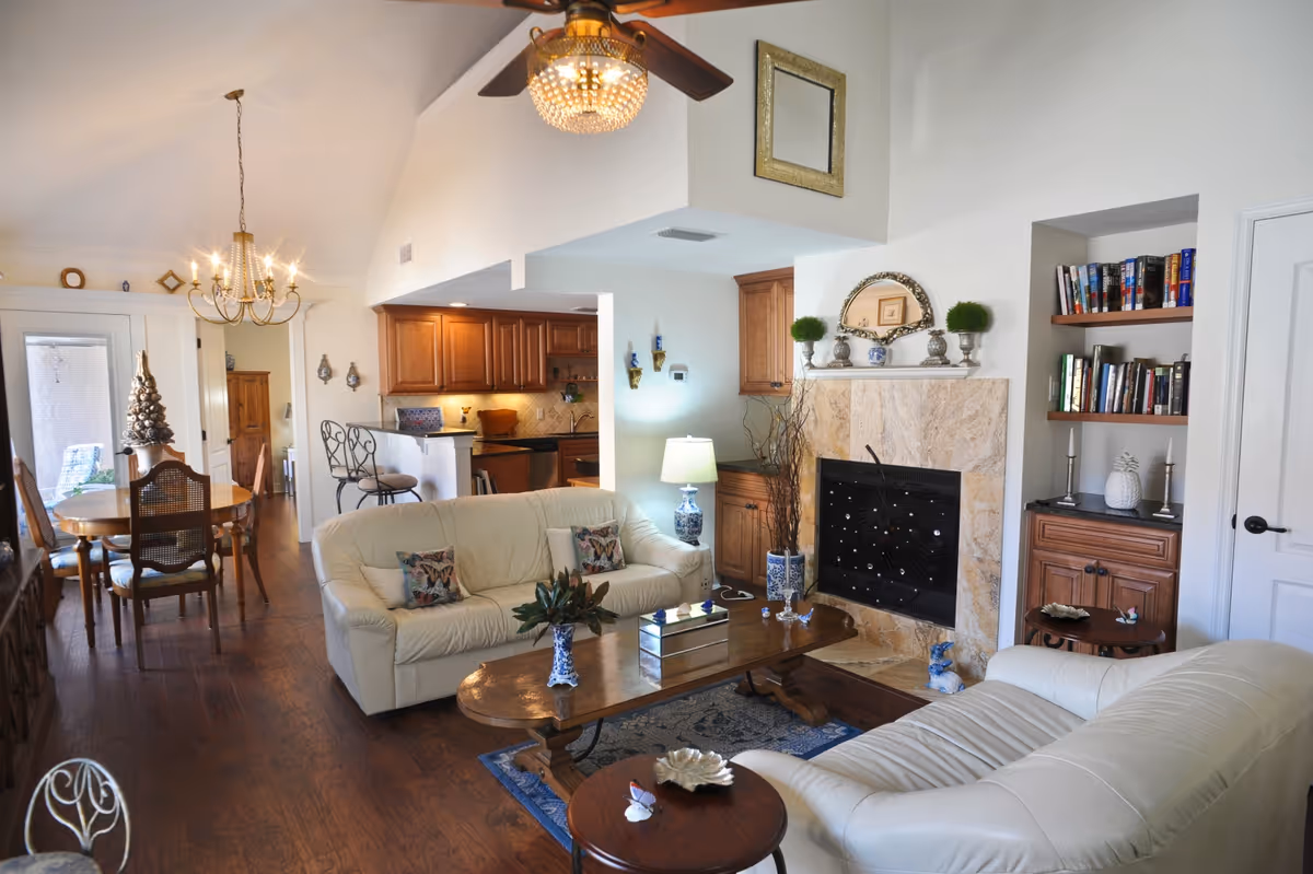 A cozy living room and dining area with cream-colored leather sofas, a wooden coffee table, and a fireplace with a decorative screen. The room features wooden flooring, built-in bookshelves, and a ceiling fan with a light fixture. The dining area has a wooden table with chairs and a chandelier overhead. The kitchen with wooden cabinets is visible in the background.