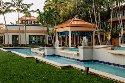 Outdoor view of Hazel Cypen Tower featuring a landscaped garden with green grass, palm trees, and a decorative water fountain with cascading water. The building has large windows and a covered patio area with a tiled roof.