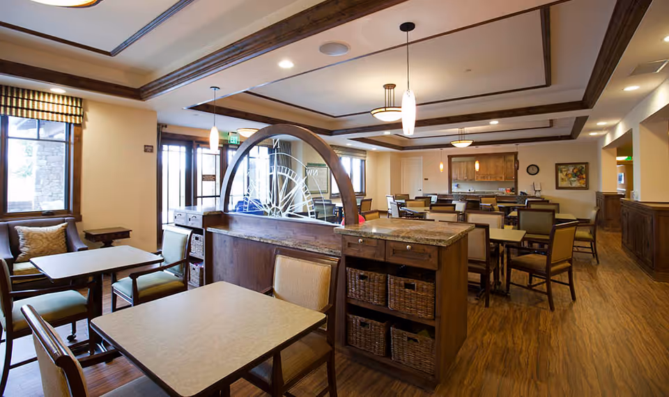 A spacious dining room in a senior living facility with multiple tables and chairs arranged neatly. The room features warm wood flooring, a decorative wooden divider with a glass design, large windows with striped valances, and soft ceiling lighting. There is a kitchen area visible in the background with wooden cabinetry and a countertop.