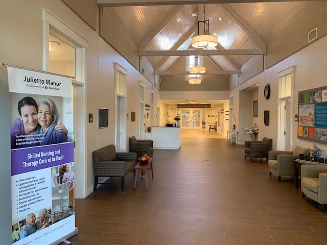 Interior view of a senior living facility hallway with high vaulted ceilings and hanging light fixtures. There are chairs and small tables arranged along the walls. On the left side, there is a standing banner for Juliette Manor advertising skilled nursing and therapy care. The hallway leads to a reception desk and glass doors at the far end.
