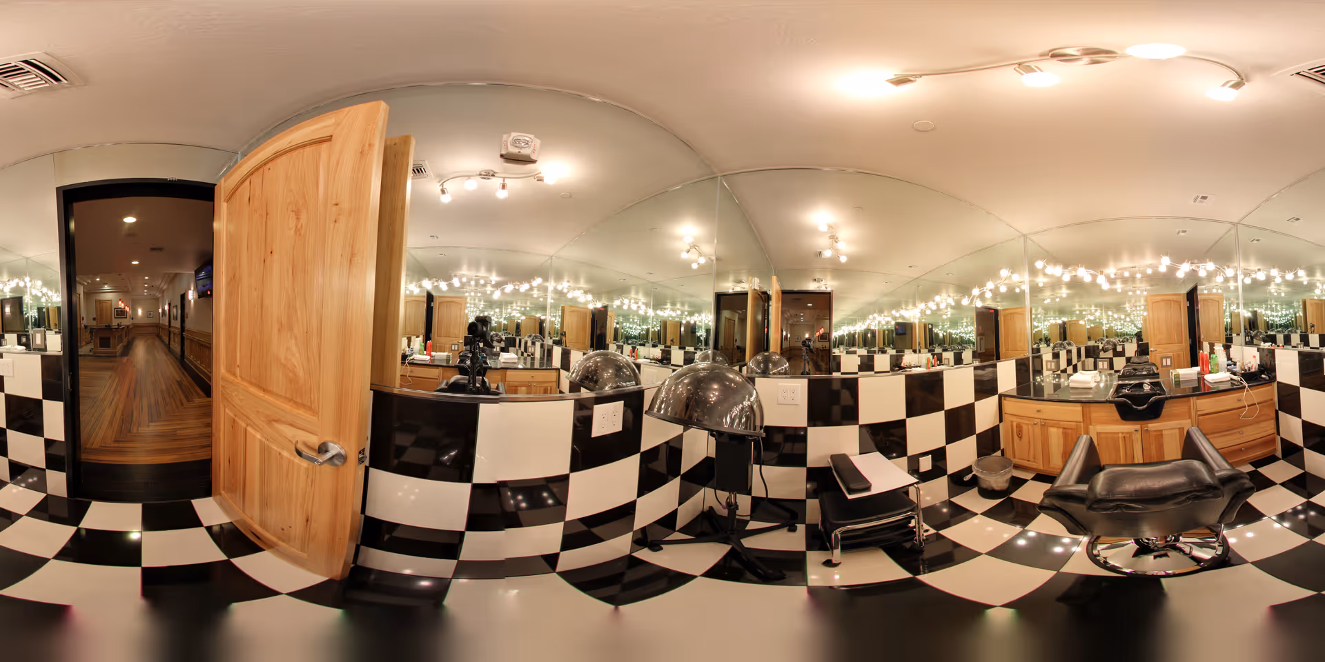 A salon area inside Heritage Creek Assisted Living featuring black and white checkered floor and wall tiles, a wooden door partially open leading to a hallway with wooden flooring, a black salon chair, a hair dryer hood, and a large mirror reflecting multiple light fixtures.