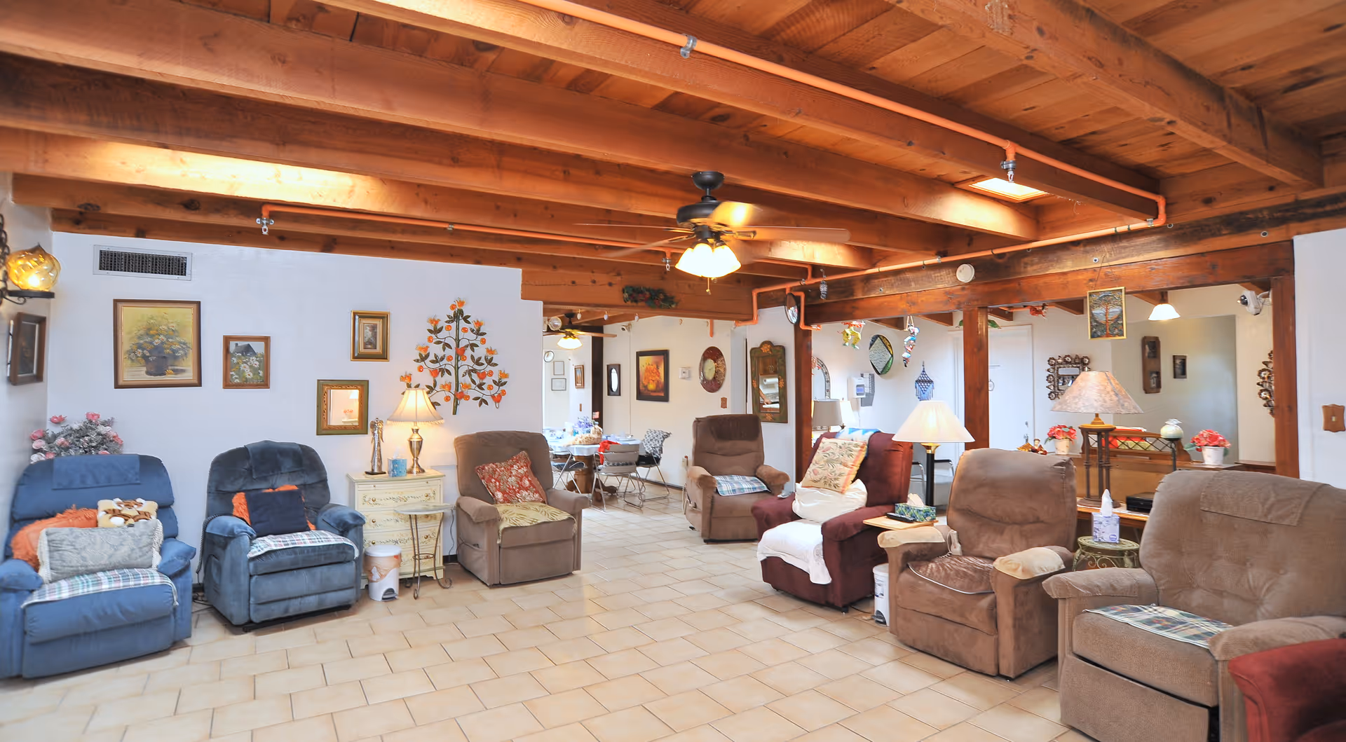 A cozy living room area with multiple recliner chairs arranged along the walls. The room features a wooden beam ceiling with a ceiling fan and several lamps providing warm lighting. The walls are decorated with framed pictures and a floral wall decal. There is a tiled floor and a small table with a lamp between some of the chairs. The space extends into a dining area in the background.