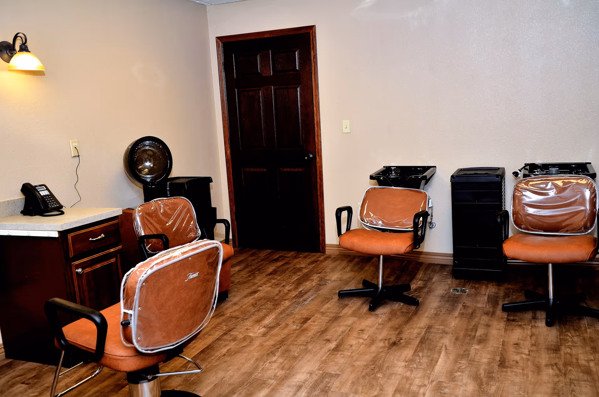 Interior of a salon area with three brown salon chairs covered in plastic, two black hair washing sinks, a telephone on a countertop, and a dark wooden door in the background. The floor is wooden and the walls are beige.