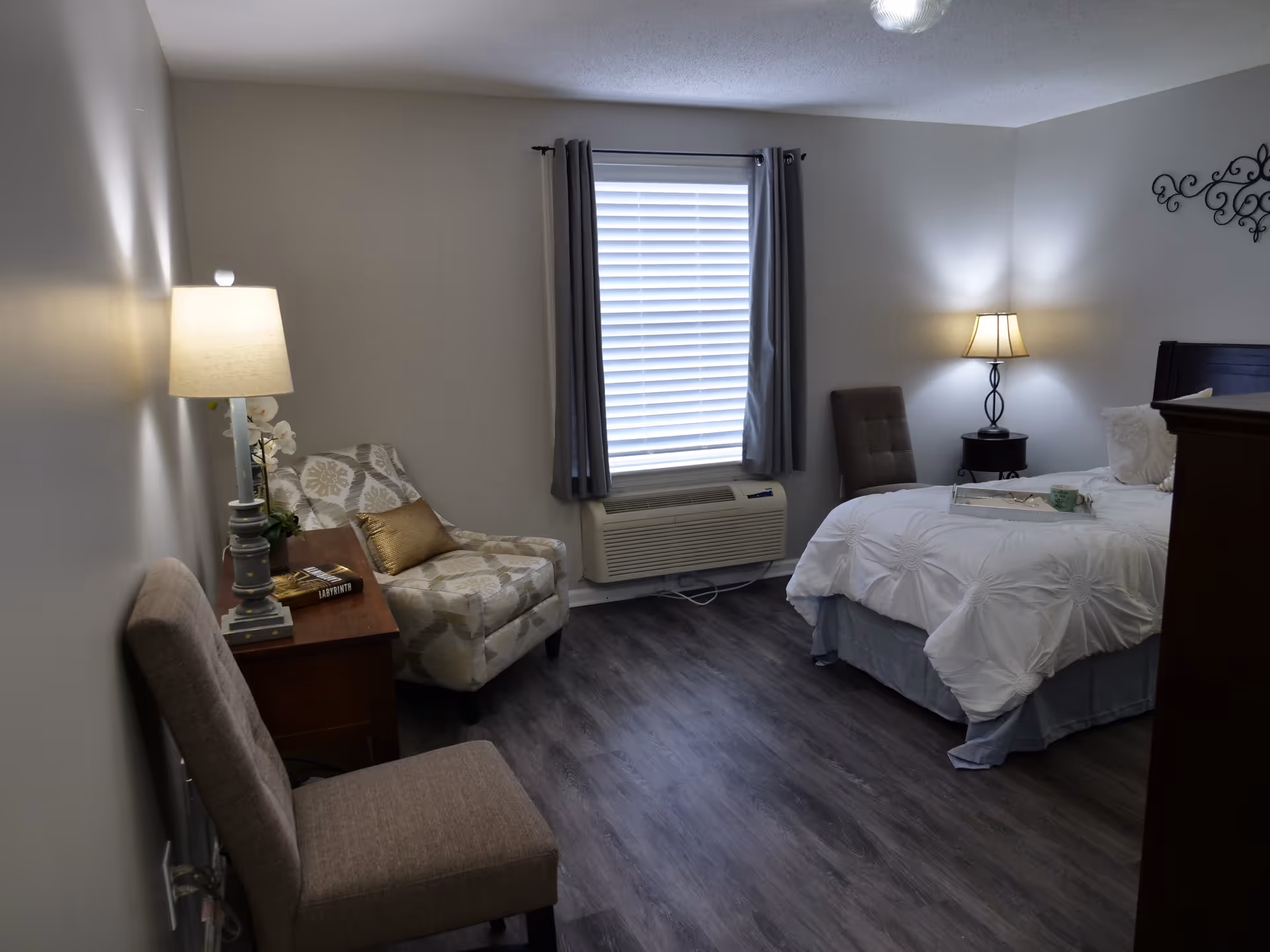 A cozy bedroom with a bed covered in white bedding, a cushioned chair, a patterned armchair, two bedside tables with lamps, a window with closed blinds and gray curtains, and dark wood flooring.