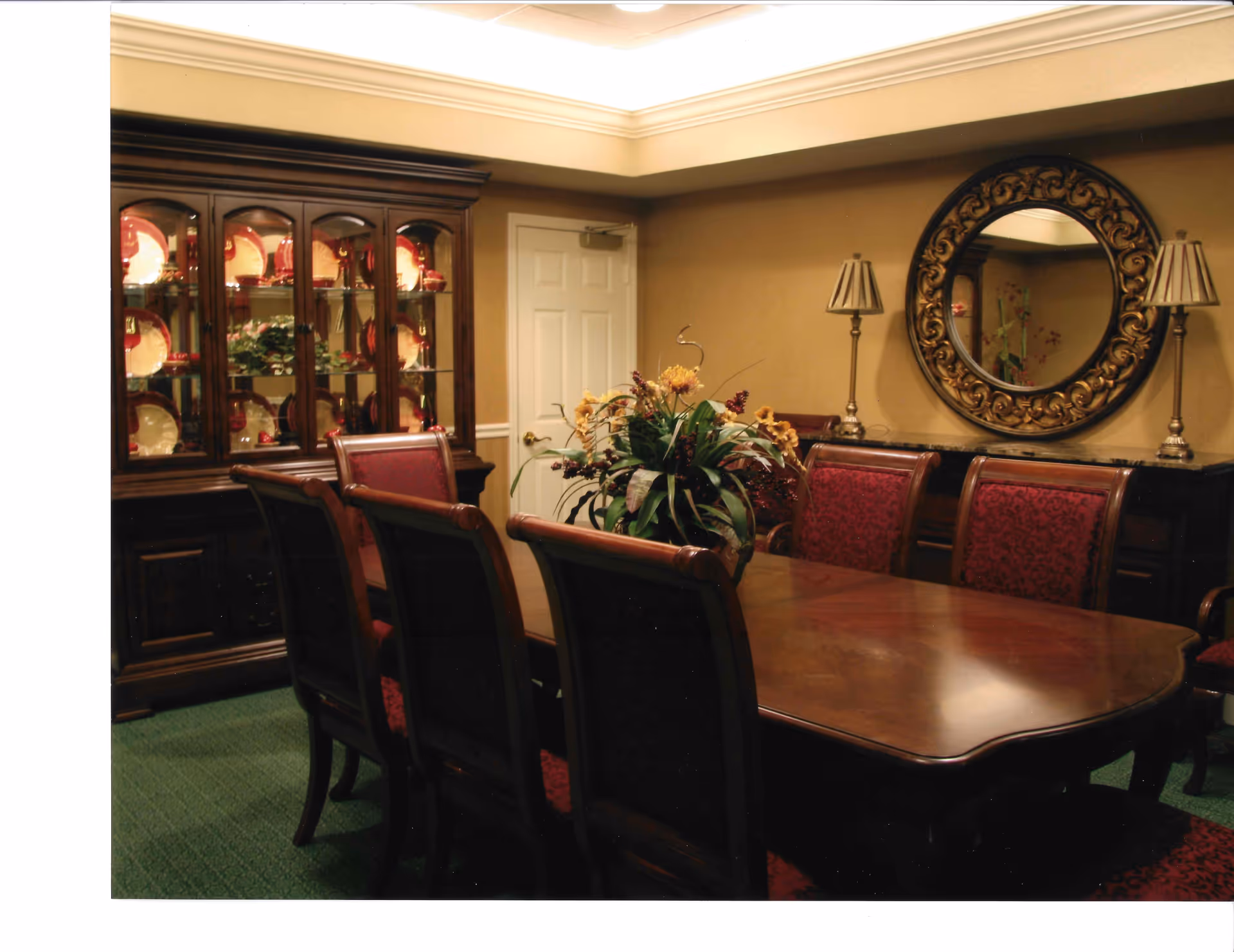 A formal dining room with a large wooden dining table surrounded by eight chairs upholstered in red patterned fabric. A floral centerpiece is placed on the table. Against the wall, there is a dark wooden china cabinet displaying red and cream-colored plates and dishes. A round ornate mirror hangs on the wall above a sideboard with two matching lamps on either side. The room has beige walls and green carpeted flooring.