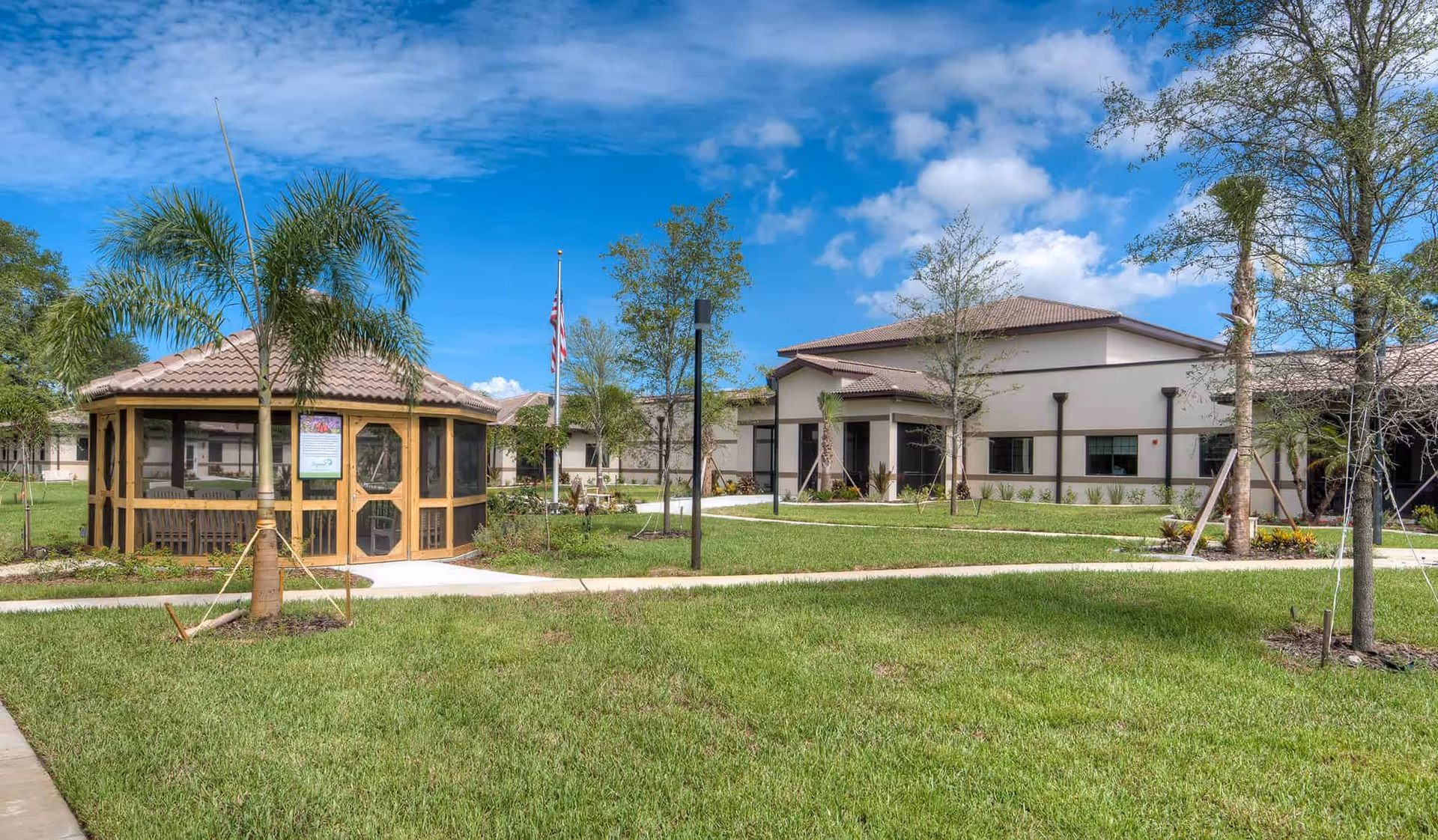 Outdoor view of Inspired Living Tampa facility showing a well-maintained lawn with young palm trees and other trees, a wooden gazebo with screened windows, a flagpole with an American flag, and a beige building with a tiled roof under a partly cloudy blue sky.