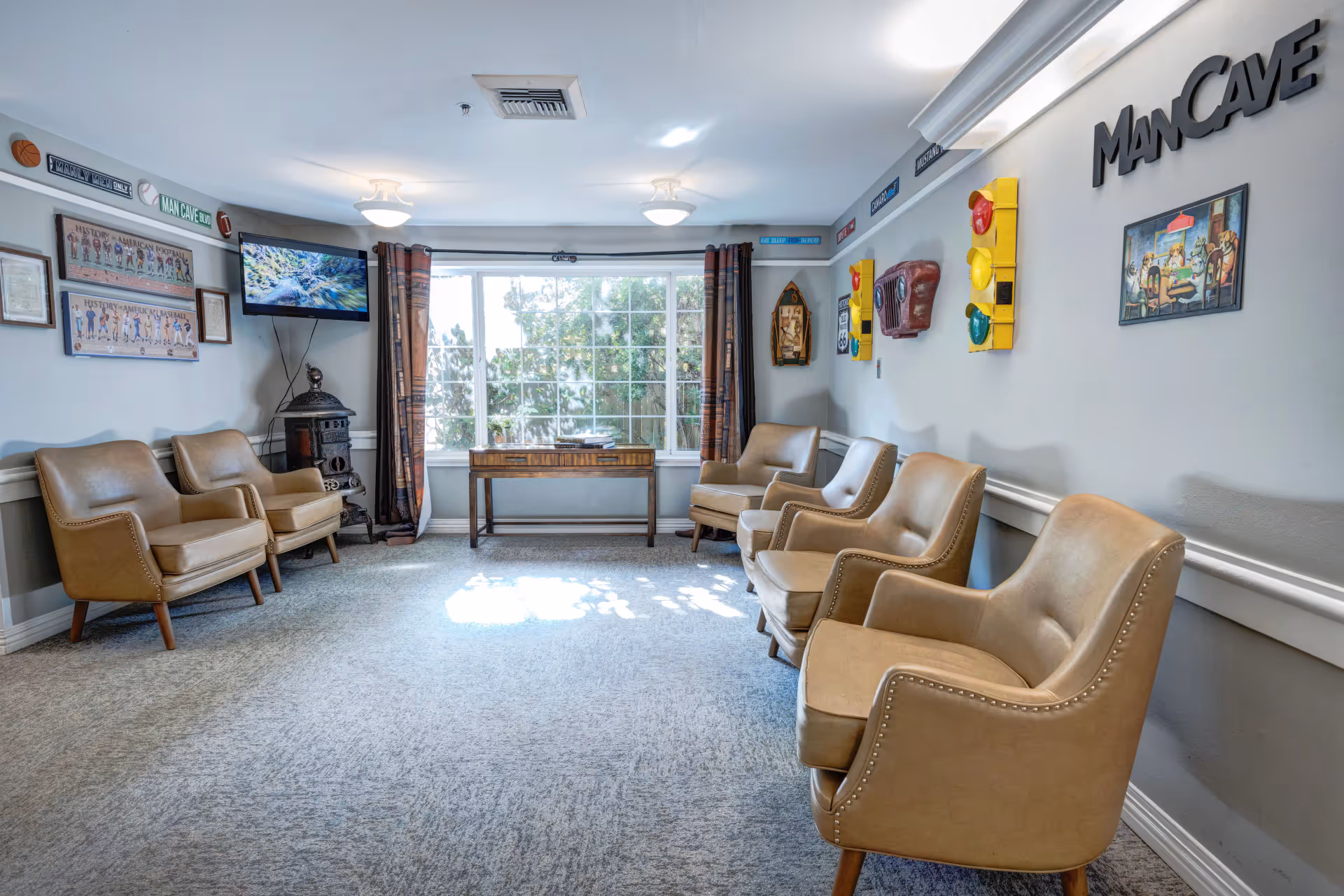 A bright seating lounge with six tan armchairs arranged along the walls, a wall-mounted TV, a large window, and a 'ManCave' sign on the wall.