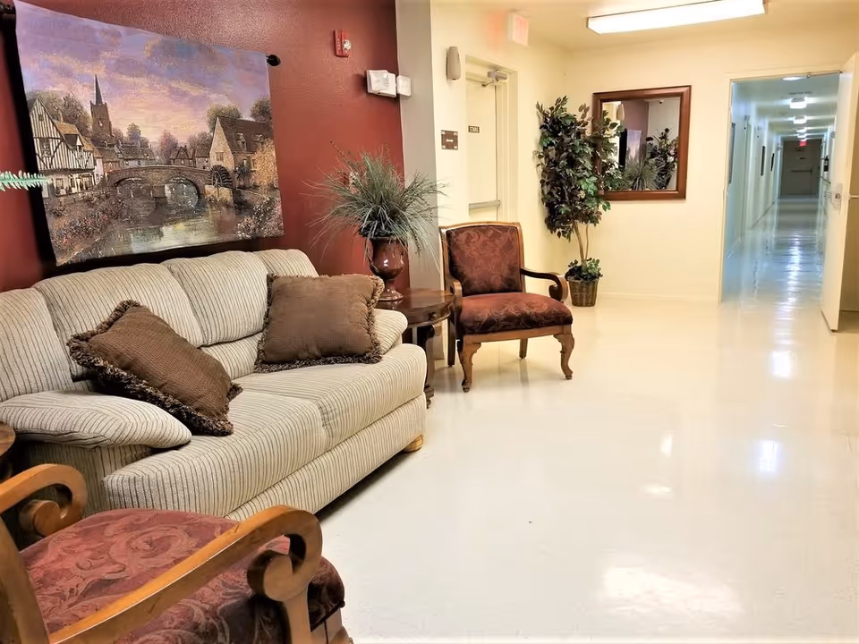 A hallway seating area in a senior living facility with a beige striped sofa adorned with brown pillows, two upholstered armchairs with wooden frames, a side table with a decorative plant, a large framed painting of a village scene on a red accent wall, a mirror on the opposite wall, and a long corridor extending into the distance.