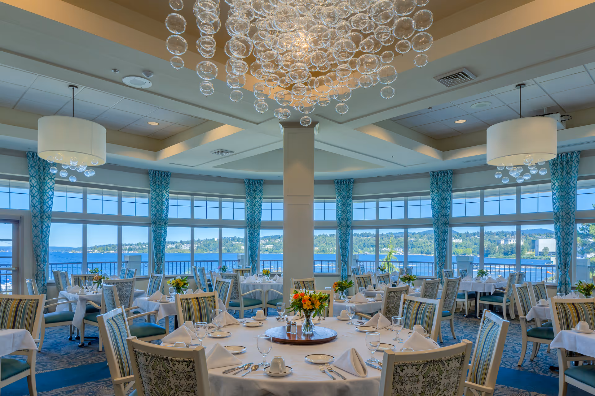 A bright and elegant dining room with round tables covered in white tablecloths, set with plates, glasses, and napkins. Each table has a centerpiece with colorful flowers. Large windows with blue patterned curtains offer a scenic view of a lake and greenery outside. Modern chandeliers with hanging glass orbs and white lampshades illuminate the room.