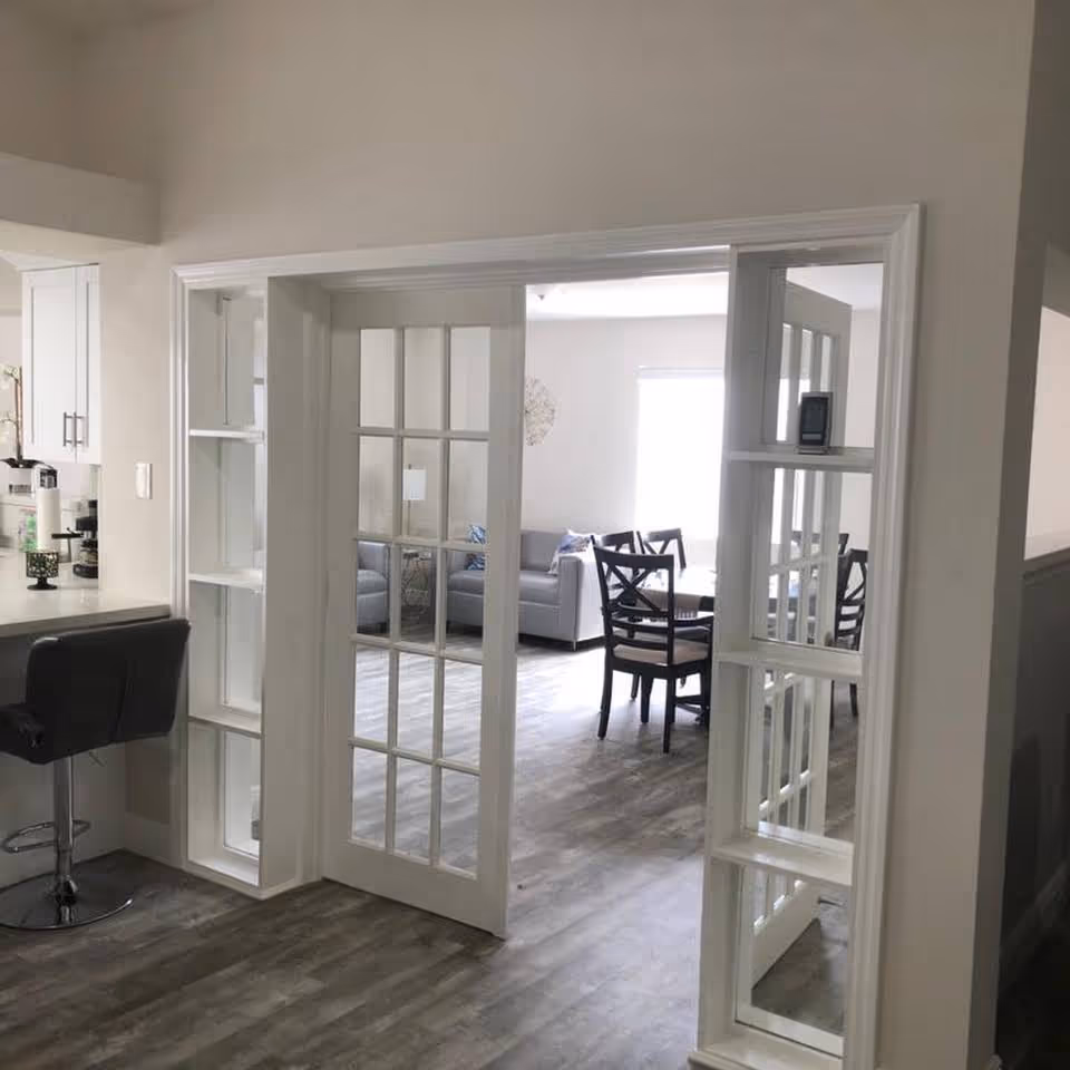 Open-plan interior showing a living and dining area through white framed glass sliding doors, with a sofa, dining table and chairs, and a bar stool at a counter.