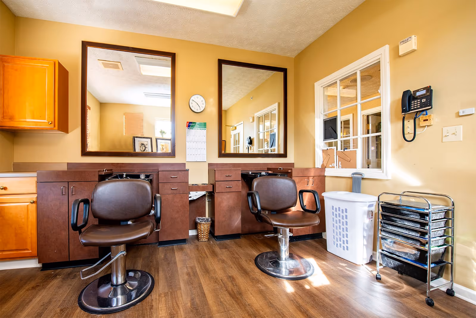 Interior view of a salon area in a senior living facility with two brown salon chairs in front of large mirrors mounted on a beige wall. There are wooden cabinets and drawers beneath the mirrors, a clock and calendar on the wall, a laundry basket, and a metal cart with trays on the right side. The floor is wooden and the room is well-lit with natural light coming through a window.