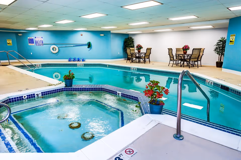 Indoor swimming pool area with a hot tub in the foreground. The pool has clear blue water and is surrounded by a beige tiled deck. There are two sets of metal handrails for entering the pool and hot tub. On the far side, there are two round tables with four chairs each, decorated with flower pots. The walls are painted blue and white, and there are safety signs and pool equipment mounted on the walls.