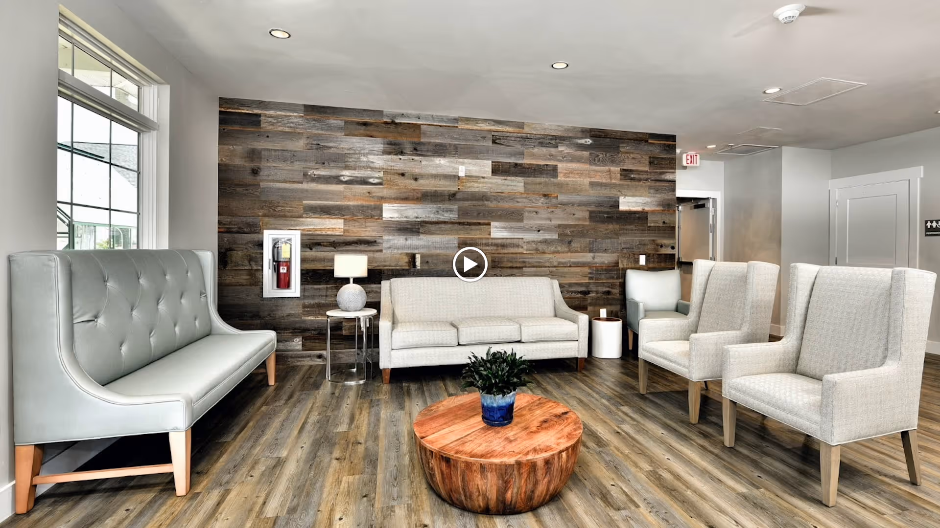 A modern living room area with a wooden plank accent wall, light gray tufted bench on the left, a light gray sofa in the center, three light gray armchairs on the right, a round wooden coffee table with a small plant in a blue pot, and a side table with a lamp. The room has wood flooring and a large window on the left side.