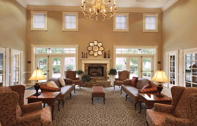 A spacious and elegant living room with high ceilings, large windows, and a central stone fireplace. The room is furnished with patterned armchairs, two long sofas, a cushioned bench, wooden side tables with lamps, and decorative plants. A chandelier hangs from the ceiling, and a flower-shaped mirror is mounted above the fireplace.